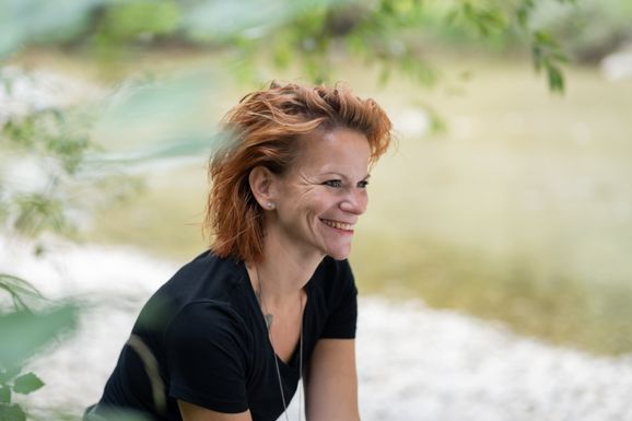 Sirga Nadja Pallotta sitzt an der Töss (Fluss) Über ihr ein Baum welcher etwas ins bild ragt. im Hintergrund sieht man das Flussbett sowie das grünliche Wasser.